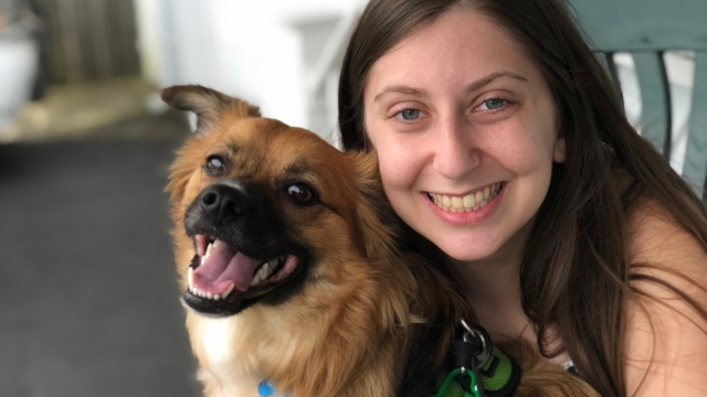 Myself-a young white woman with blue eyes and brown hair-smiling while holding my dog, a tan long-haired, small dog with a black snout.